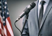 Man in suit speaking into microphone, standing next to American flag.