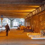 People walk under a bridge with homeless encampment