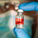 Trump’s Vaccine Ban Shakes Big Pharma Gloved hand holding vial with red liquid and flag