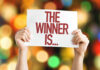Hands holding a sign reading The Winner Is