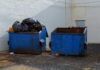 Two blue dumpsters, one overflowing with trash bags.