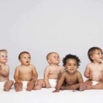 Five diverse babies sitting on a neutral background, displaying playful expressions
