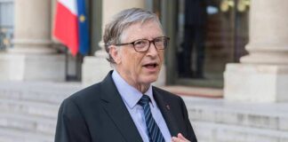 A man in formal attire speaking outdoors with a flag in the background