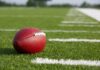 NFL Reeling After Rising Star’s Shocking Death A red American football resting on a green artificial turf field