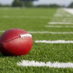 NFL Reeling After Rising Star’s Shocking Death A red American football resting on a green artificial turf field