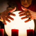 A person with adorned hands hovering over a glowing crystal ball surrounded by candles