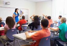 Teacher in a blue dress instructing students in a classroom with hands raised