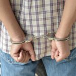 A person in handcuffs viewed from behind, wearing a plaid shirt and denim jeans