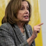 Woman speaking and pointing, wearing a plaid jacket.