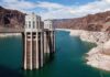 A large dam structure with two towers over a blue water reservoir surrounded by rocky mountains