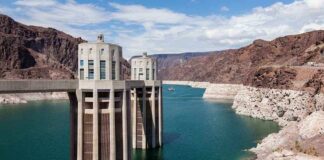 A large dam structure with two towers over a blue water reservoir surrounded by rocky mountains