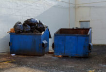 Two blue dumpsters, one overflowing with trash bags.