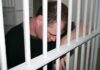 A man sitting in a jail cell with his head down, resting on the bars