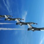 Four military jets flying in formation against a blue sky, leaving smoke trails