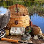 Fishing gear including a hat, basket, and fishing rods arranged by a serene water body