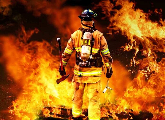 Firefighter standing in front of a large fire, equipped with tools