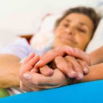 A caregiver holding the hands of an elderly patient in a hospital bed