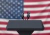 Ex-Researcher Claims ‘Four Species’ Are Here! Podium with microphones, American flag background.