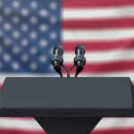 Ex-Researcher Claims ‘Four Species’ Are Here! Podium with microphones, American flag background.