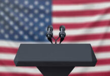 Podium with microphones, American flag background.