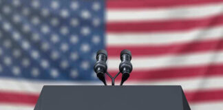 Podium with microphones, American flag background.