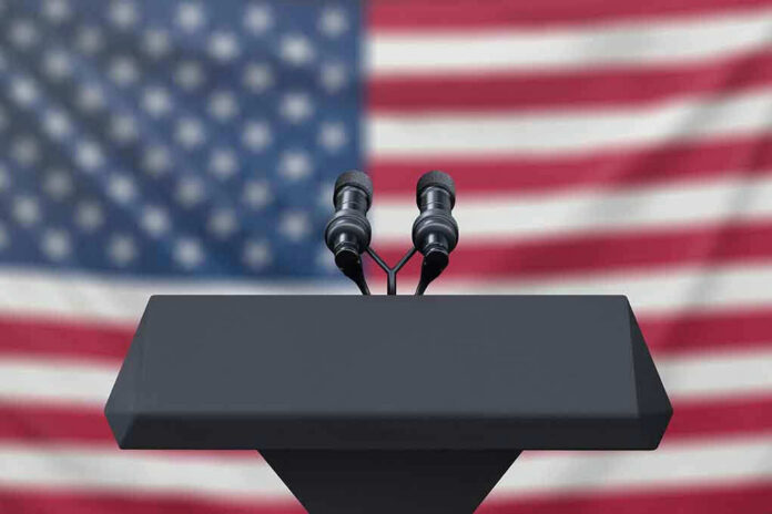 Podium with microphones, American flag background.