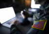 War Plans Vanish In ‘Auto-Delete’ Backchannel Soldier using laptop with US flag patch visible.