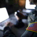 War Plans Vanish In ‘Auto-Delete’ Backchannel Soldier using laptop with US flag patch visible.