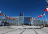 Finland Ends Its Nuclear Ban To Plug Into NATO’s Deterrence Flags outside NATO headquarters building under clear blue sky.