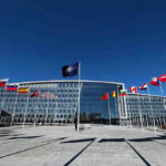 Finland Ends Its Nuclear Ban To Plug Into NATO’s Deterrence Flags outside NATO headquarters building under clear blue sky.