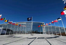 Finland Ends Its Nuclear Ban To Plug Into NATO’s Deterrence Flags outside NATO headquarters building under clear blue sky.