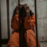 Idaho’s Firing Squad: Quick Death Or Torture? Person in orange jumpsuit sitting behind bars, handcuffed.