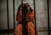 Person in orange jumpsuit sitting behind bars, handcuffed.