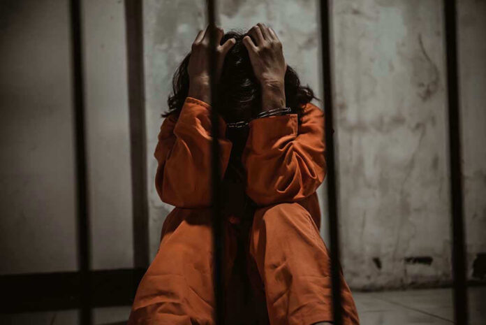 1484320151 Person in orange jumpsuit sitting behind bars, handcuffed.