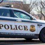 Agent Googles As Bullets Fly United States Secret Service police car on roadside.