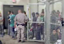 Detention facility with people behind chain-link fence and officer nearby