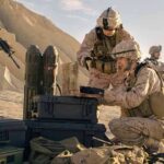 Soldiers in desert setting prepare equipment near rugged hills