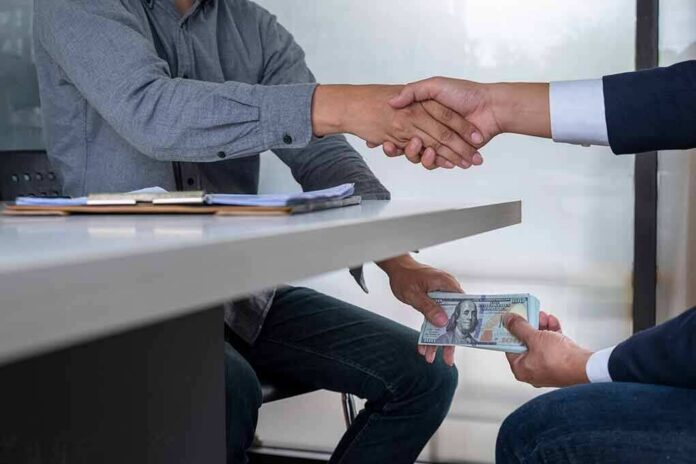 shutterstock_1169658241 Two people shake hands over a desk while cash is exchanged underneath