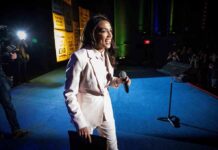 AOC’s Gala Dress Maker Ends in a Seven-Figure Lawsuit Woman in a light suit holding a microphone on stage