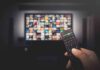 Hand holding TV remote toward a screen with streaming thumbnails