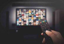 Hand holding TV remote toward a screen with streaming thumbnails