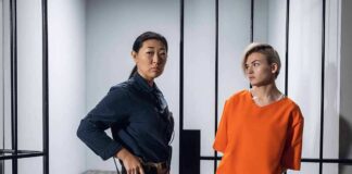 Police officer stands beside inmate in orange uniform inside a jail cell