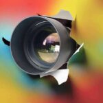 camera lens emerging through torn paper on colorful background