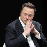 Elon Musk seated in a dark setting with hands steepled near his face